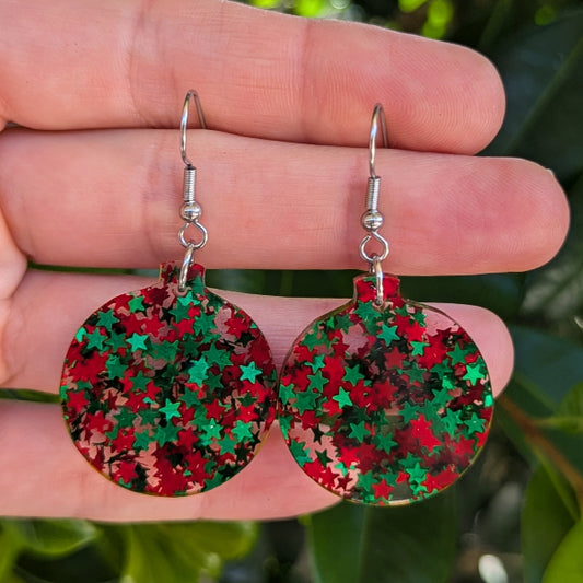 A pair of round silver-tone dangle earrings covered in red and green star-shaped glitter, held between a person's fingers against a blurred green outdoor background.