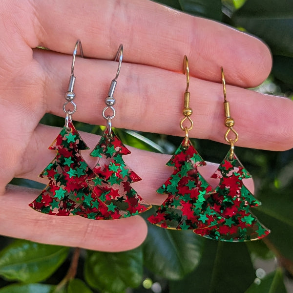 Two pairs of dangling earrings shaped like Christmas trees, decorated with red and green glitter stars, held in a hand against a background of green leaves. The hooks are silver-tone on the left, gold-tone on the right.