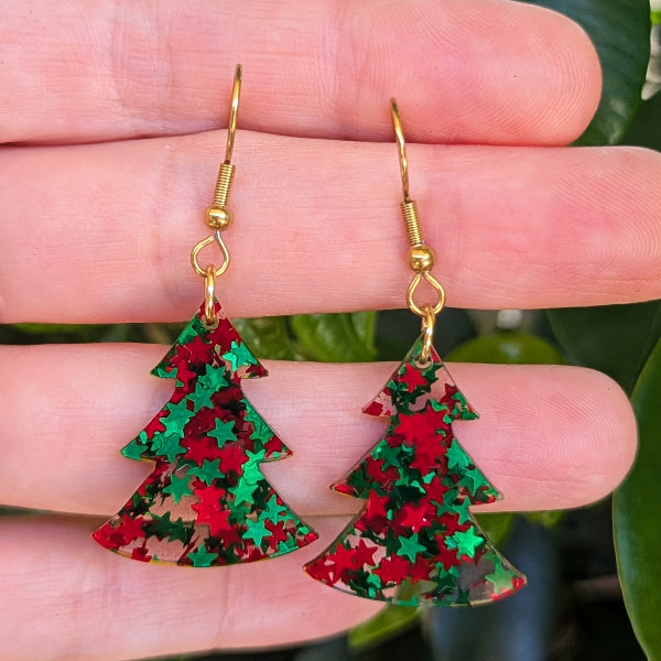 A pair of dangling earrings shaped like Christmas trees, covered in red and green glitter, hanging from gold fishhook wires and held against a person's fingers with green leaves in the background.