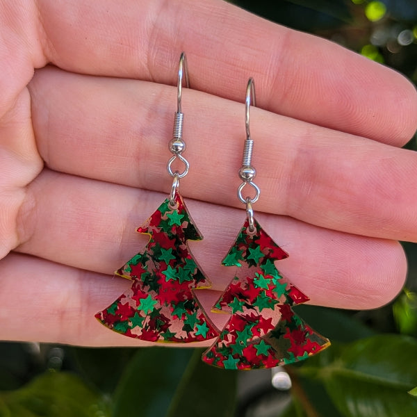 A pair of dangling earrings shaped like Christmas trees, filled with red and green glitter, hanging from silver fishhook wires. They are held in a person's hand against a blurred background of green leaves.