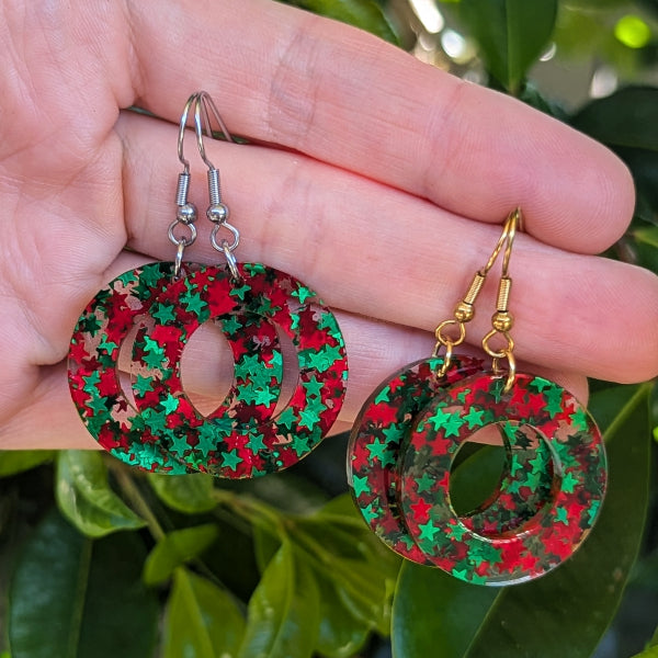 A pair of round double-ring dangle earrings with a red and green star pattern, shown hanging from a person's fingers with green leaves in the background.