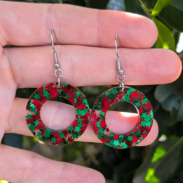 A pair of silver-tone dangle earrings with circular cutouts filled with red and green star-shaped glitter, held on a person's hand with blurred green foliage in the background.
