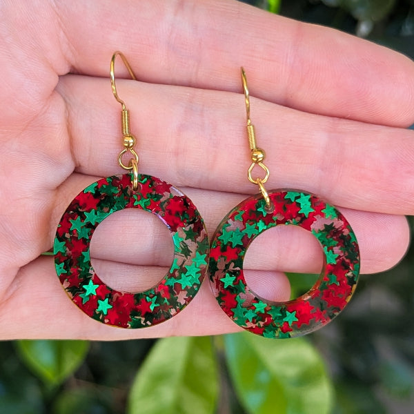 A pair of gold-tone dangle earrings with circular cutouts filled with red and green star-shaped glitter, held in a person's hand against a blurred green outdoor background.