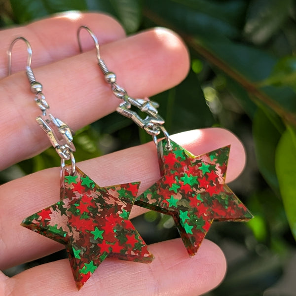 A pair of dangling star-shaped earrings with a red and green glittery pattern, held in a person's hand against a blurred background of green leaves.