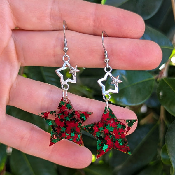 A pair of dangling star earrings held in a hand, featuring red and green glitter-filled star pendants with silver-tone metal hooks and star charms, against a background of green leaves.
