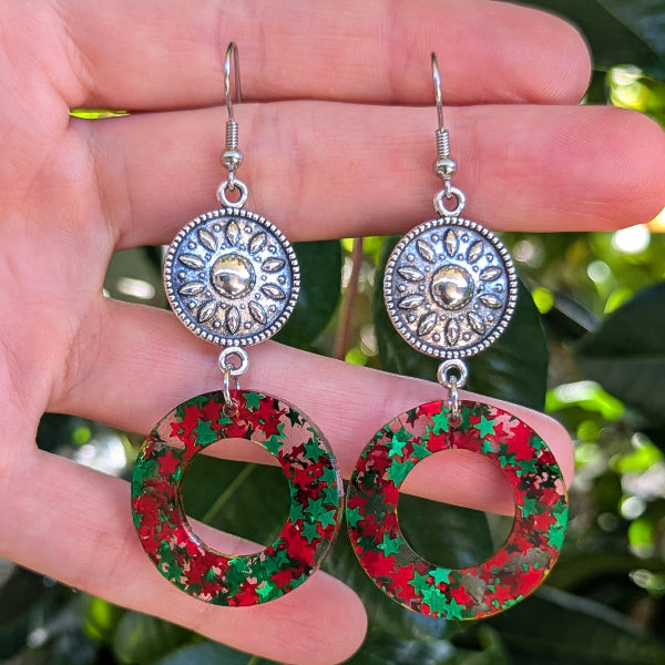A pair of dangling earrings held in a hand, featuring silver-tone circular filigree tops and red-and-green glittery ring drops, with blurred green foliage in the background.