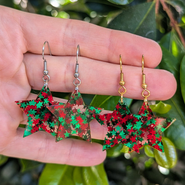 Four star-shaped dangling earrings with red and green glittery panels are displayed on a person's hand, with green foliage in the background.