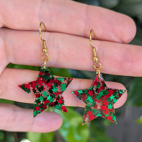 A pair of dangling star-shaped earrings with gold fishhook wires and a red and green star-patterned glitter face, shown hanging from a person's fingers against a blurred green background.