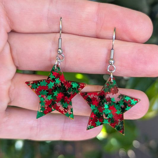 A pair of star-shaped dangling earrings covered in red and green star confetti, held in a person's hand against a blurred green outdoor background.