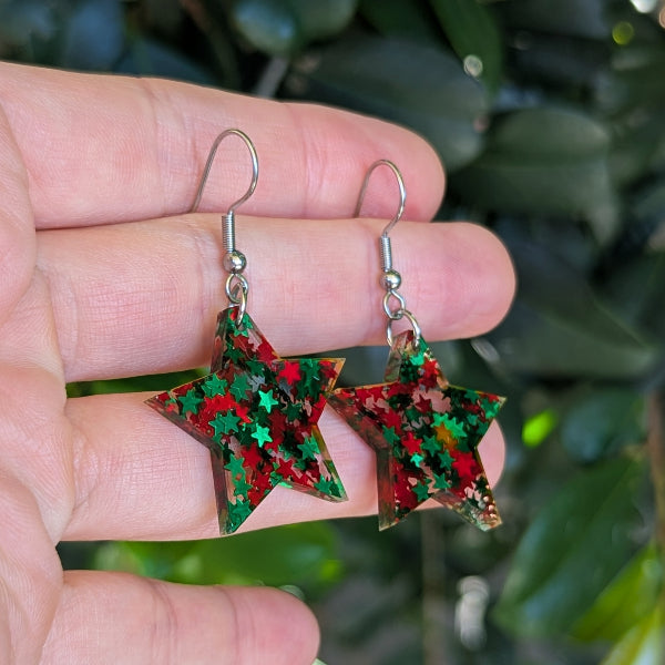 A pair of star-shaped dangling earrings with red and green glitter, held in a person's hand against a blurred background of green leaves.