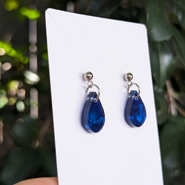 A pair of deep blue teardrop-shaped drop earrings with silver-toned posts and rings, displayed on a white card against a blurred green outdoor background.