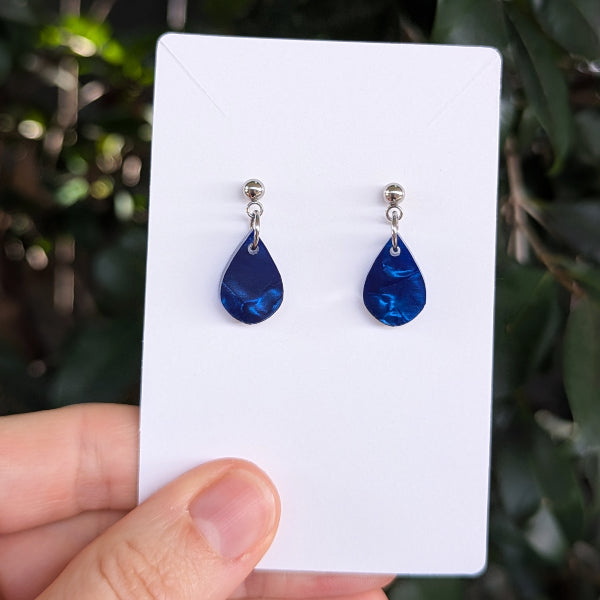 A pair of deep blue teardrop-shaped drop earrings with silver posts, displayed on a white card held by a hand against a blurred green outdoor background.