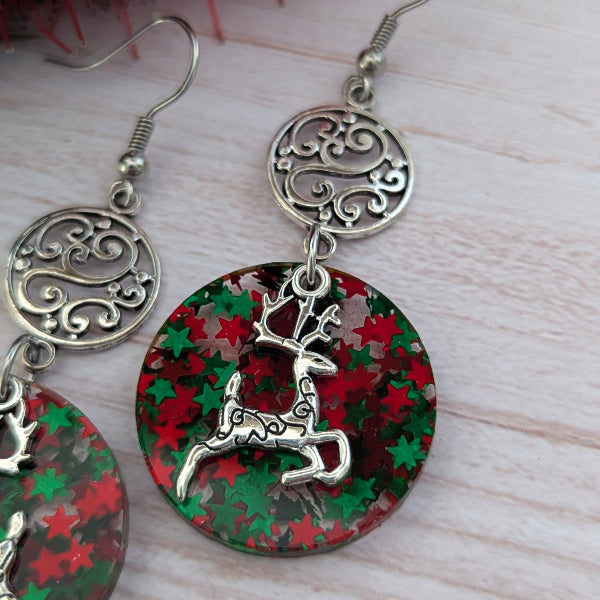 Christmas-themed dangle earrings with ornate silver filigree circular tops, green and red star-patterned round drops, and silver reindeer charms, shown on a light wood background.