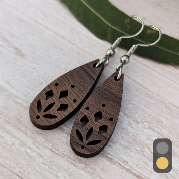 A pair of teardrop-shaped wooden dangle earrings with laser-cut leaf patterns, featuring dark brown wood grain and silver-tone fishhook ear wires, displayed on a light wood surface with a green leaf prop.