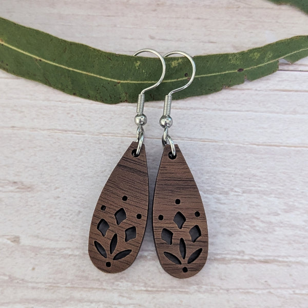 A pair of teardrop-shaped wooden dangle earrings with laser-cut floral patterns, hanging from silver fishhook wires. The earrings are displayed on a light-colored wooden surface with a green leaf prop.