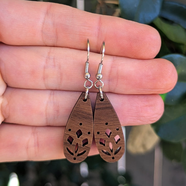 A pair of wooden teardrop dangle earrings with laser-cut leaf patterns, held in a person's hand against a blurred background of green leaves.