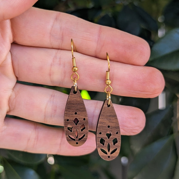 A pair of wooden teardrop dangle earrings with gold fishhook ear wires, held in a person's hand against a blurred background of green leaves.