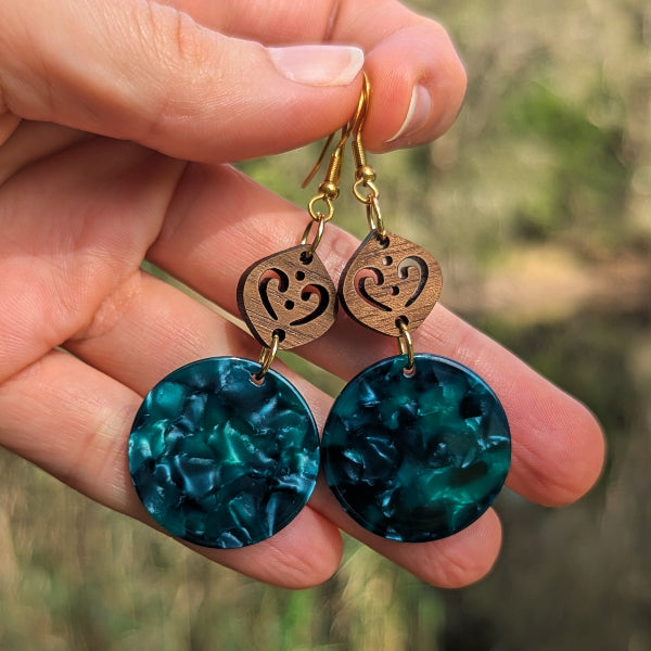 A close-up of a hand holding a pair of bohemian-style dangle earrings. The earrings feature round, marbled green disc pendants with gold-tone hooks and carved wooden charms.