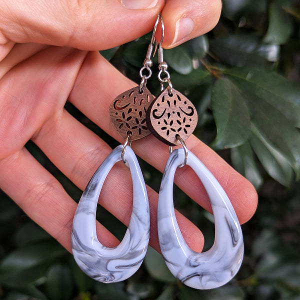 A pair of teardrop-shaped dangle earrings with white and gray marbled resin pendants and wooden filigree charms, held in a hand against a background of green leaves.