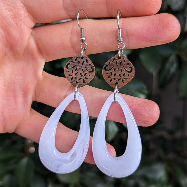 A pair of teardrop-shaped dangle earrings held in a hand. The earrings feature a silver-tone hook ear wire, a small oval wooden disc with an intricate cutout pattern, and a larger translucent white teardrop drop.